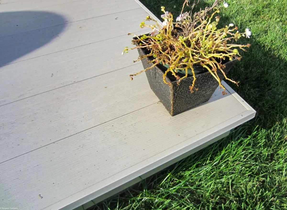 A black square planter with a decorative pattern, containing dried and wilted pink and white flowers, sits on a white outdoor deck beside green grass.