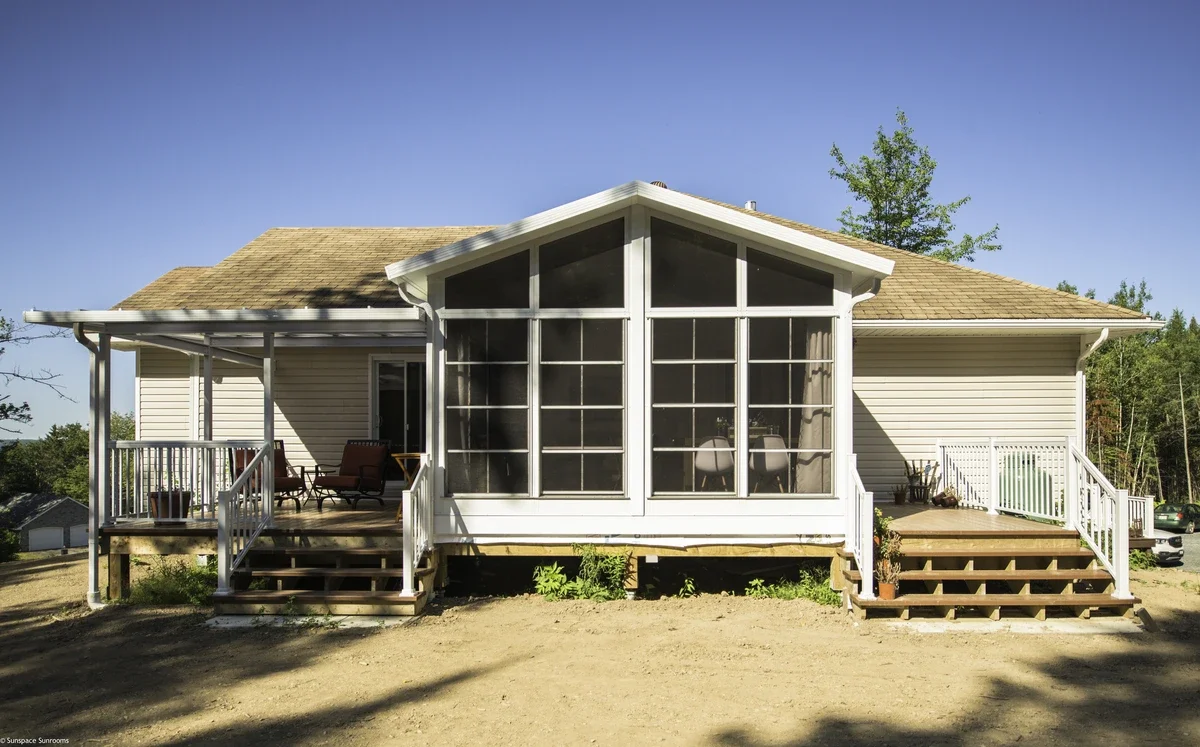 Rear view of a house with a screened porch and two stairs leading up to it, surrounded by a dirt yard and trees in the background.