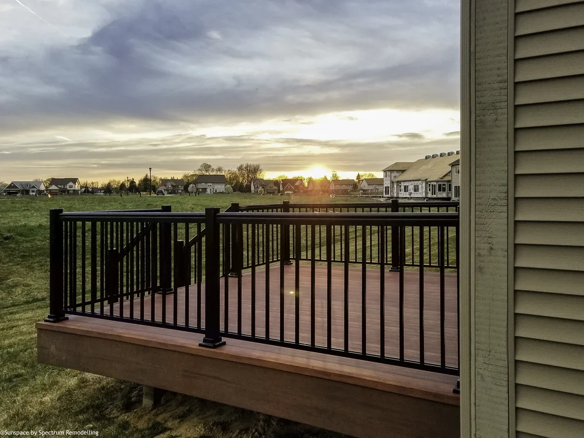 A wooden deck attached to the side of a house with a black metal railing, overlooking a grassy field and houses in the distance during sunset under a cloudy sky.