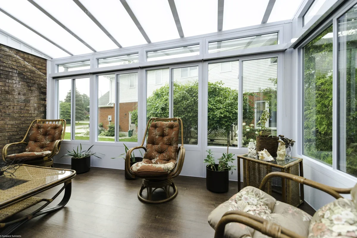 Sunroom with wicker chairs, potted plants, and decorative items, overlooking a backyard garden with trees and a house.