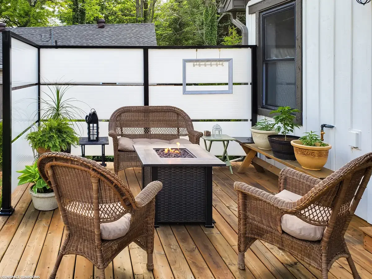 Outdoor patio with wicker chairs, a fire table, potted plants, and decorative elements on a wooden deck enclosed by a white privacy fence.
