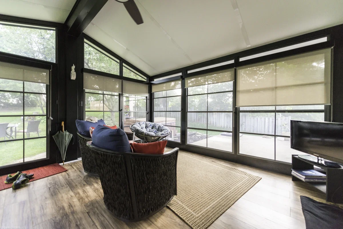 Sunroom with black window frames, beige blinds, wooden floor, wicker chairs with cushions, a small black TV on a stand, and an outdoor view of grass and trees.