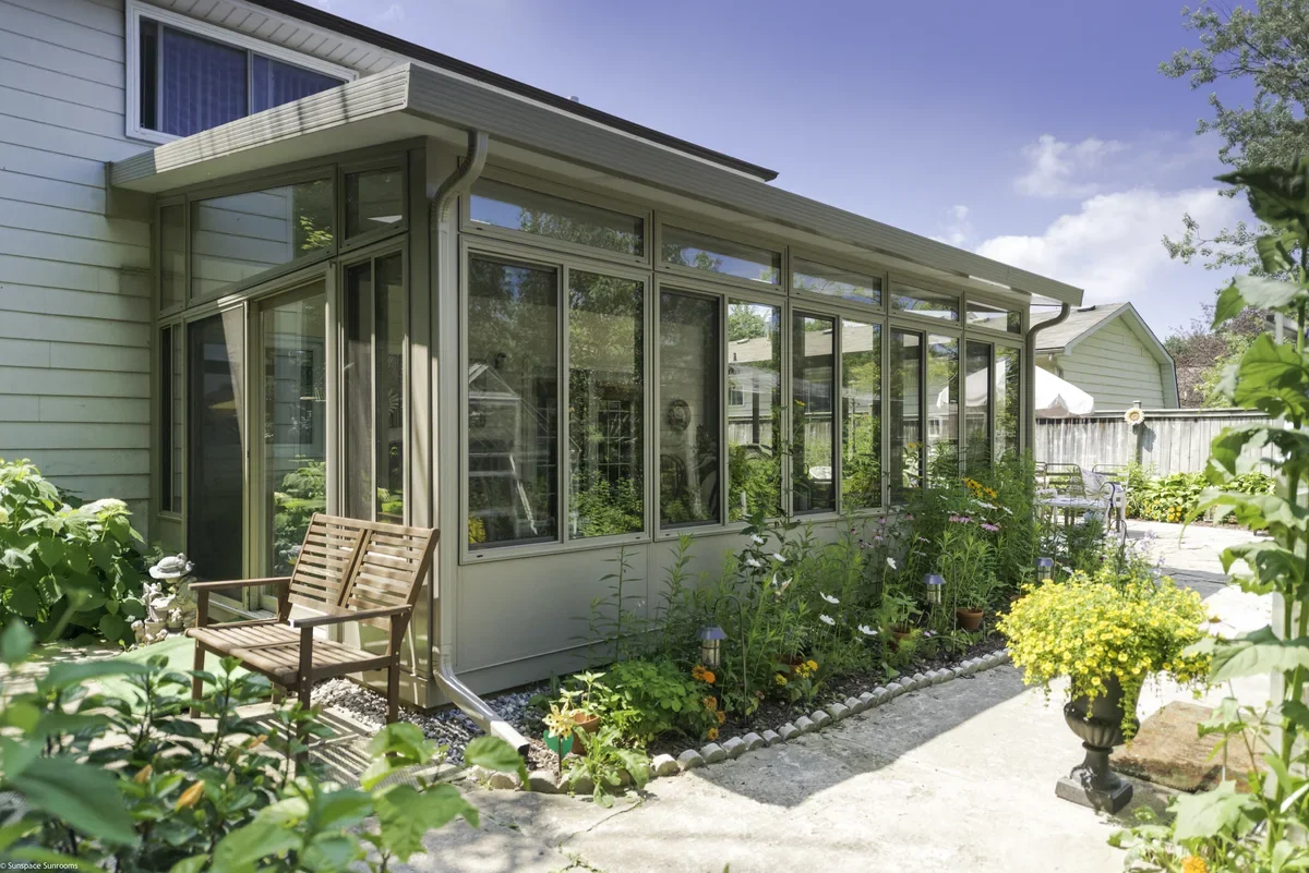 A backyard garden with a sunroom attached to a house, green plants, yellow flowers in a black urn, and outdoor furniture on a patio.
