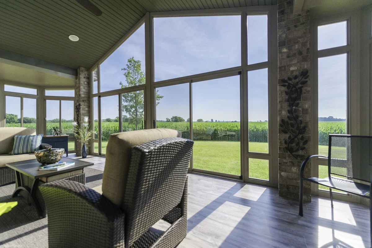 Sunroom with large windows overlooking a green field, wicker chairs, a sofa with pillows, a small table with decorative items, and a potted plant.