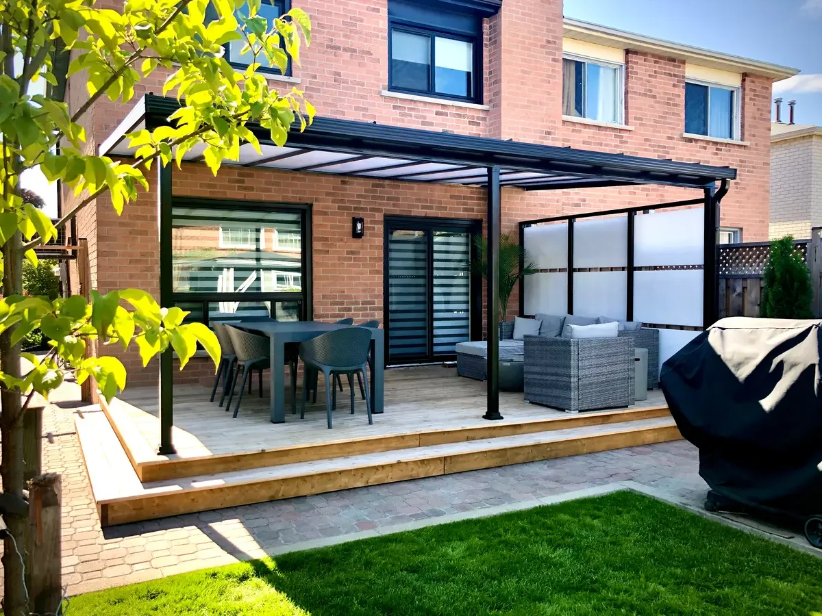A backyard patio with a wooden deck, outdoor furniture including a round table with six chairs, and a seating area with a woven sofa and matching chairs. The patio is covered with a black metal pergola with a translucent roof. There is a black grill covered with a black cover, a window, and a sliding door on the house's brick exterior. The yard has a small patch of green grass and a tree with bright green leaves.
