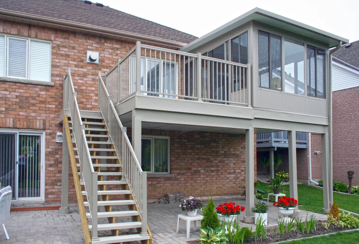 Backyard of a house with a brick house and a wooden elevated deck with stairs leading down, adorned with potted plants and garden flowers.