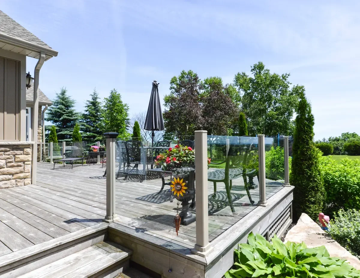An outdoor wooden deck patio with black metal tables and chairs, a closed black patio umbrella, potted flowers, and a glass railing, surrounded by green trees and shrubs under a clear blue sky.