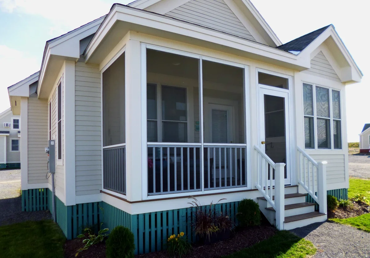 A small, modern house with a screened front porch, white siding, and a staircase leading to the porch. The house is surrounded by a simple landscape with small bushes and grass.