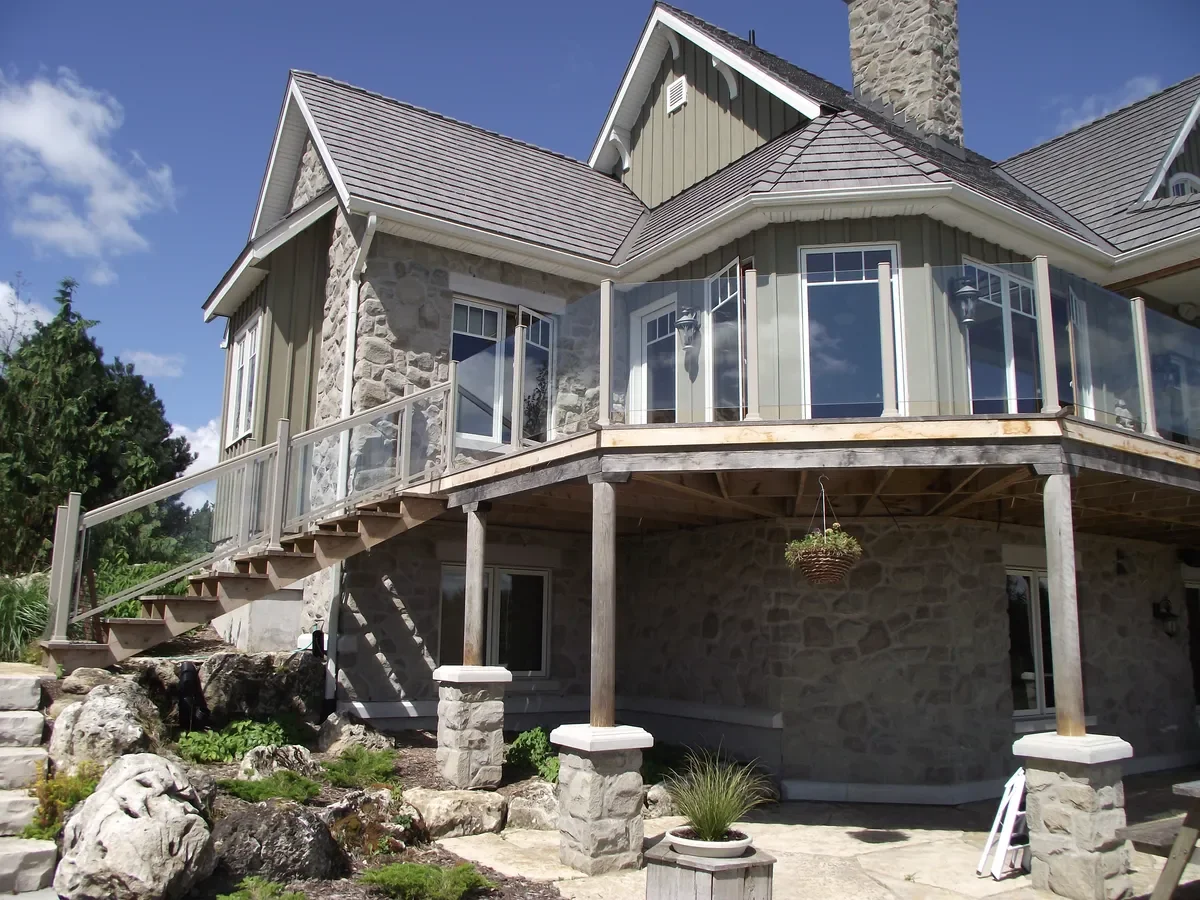 Multi-story house with a stone and siding exterior, large windows, a wrap-around deck with a glass railing, and outdoor stairs leading to the yard. The house has a shingled roof and a chimney.