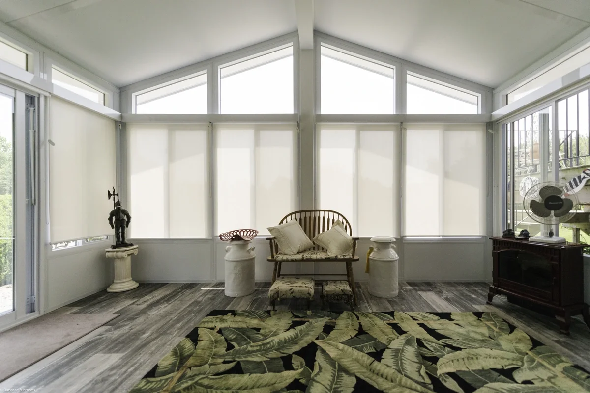 Sunroom with large windows, white blinds, a wooden bench with pillows, decorative pedestal tables, a patterned rug with tropical leaves, and a dark wooden cabinet with a fan and decorative items.