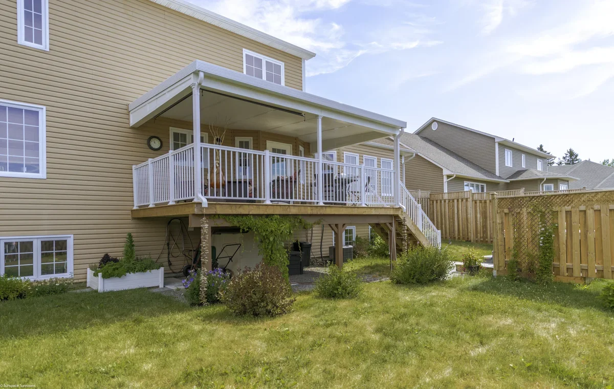 Backyard with a raised wooden deck attached to a yellow house, fenced yard, and neighboring houses in the background, under a partly cloudy sky.