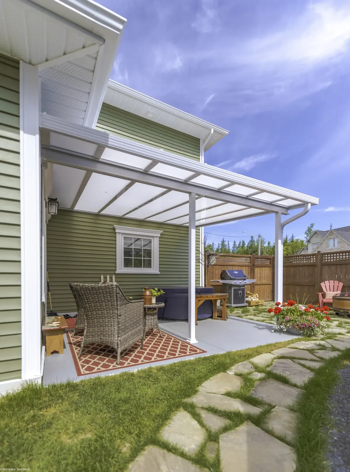 Backyard patio with a green house, white pergola, outdoor furniture, a grill, and flower beds under a blue sky.