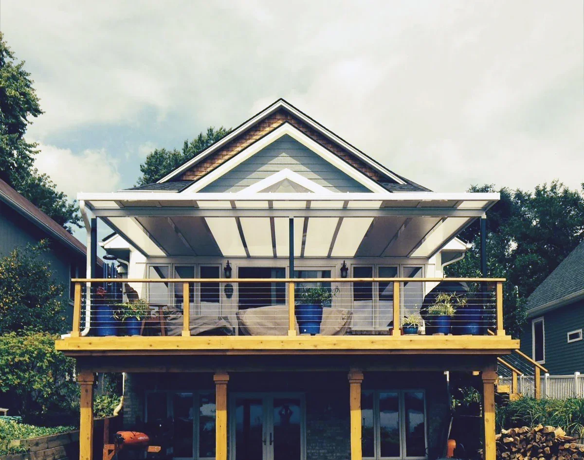 Two-story house with a large balcony on the upper floor, featuring a retractable white awning, potted plants, and outdoor seating. The house has light gray siding, a gable roof, and surrounding greenery.