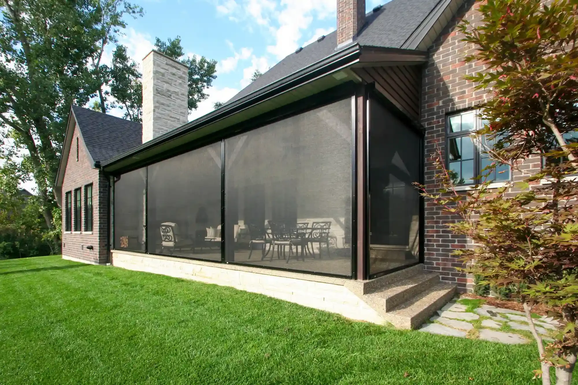 A house with brick walls and a screened-in porch with outdoor furniture, surrounded by green grass, trees, and a blue sky with clouds.