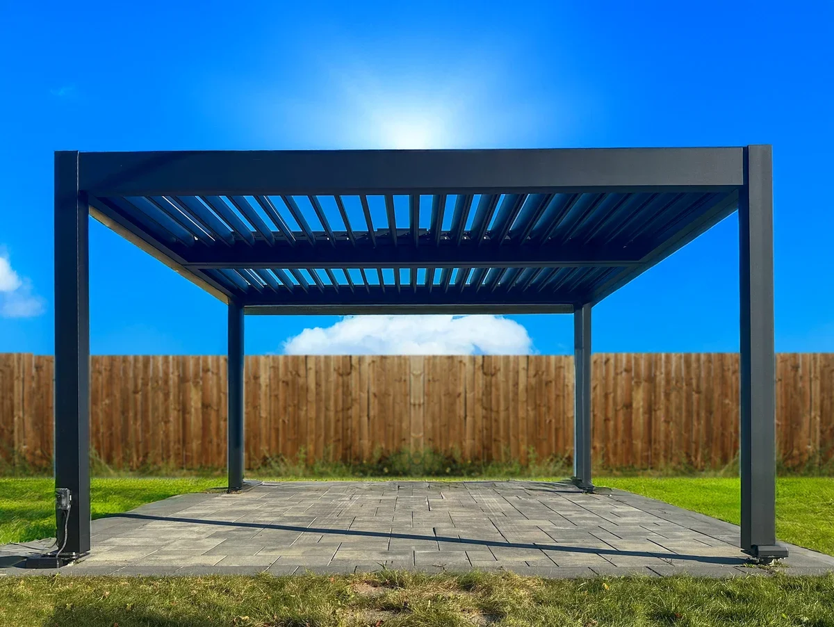 An outdoor metal pergola with a slatted roof on a paved patio, surrounded by a grassy yard and a wooden fence under a clear blue sky.