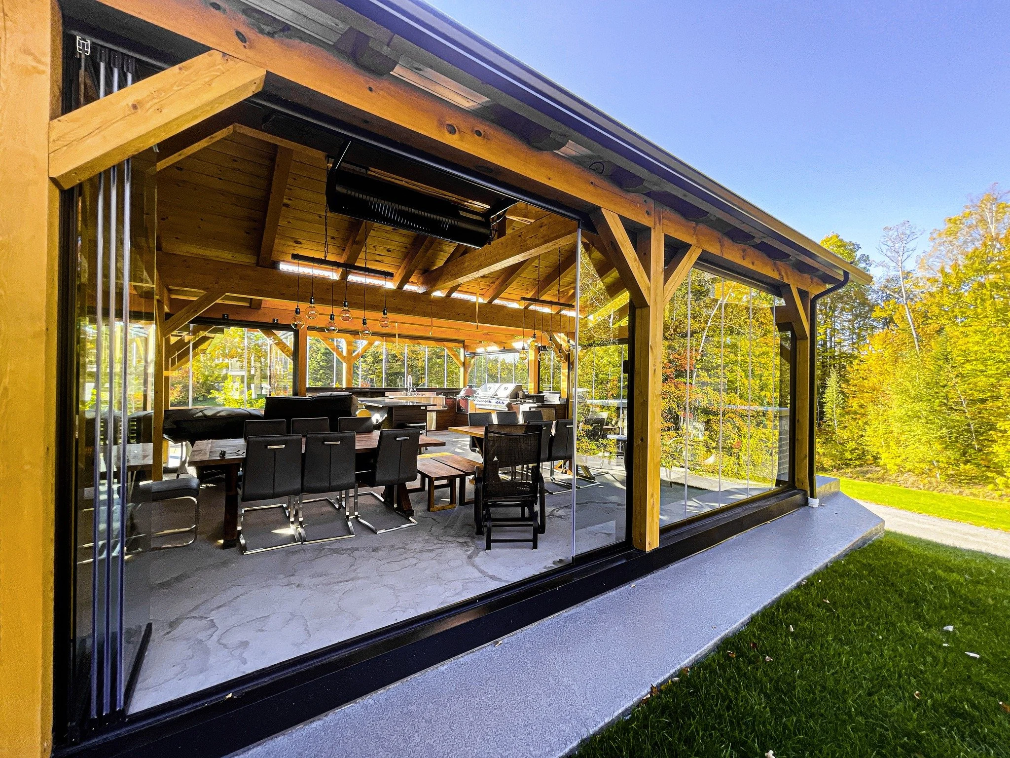 View of a modern house with large glass windows, a wooden roof support structure, and an outdoor patio with seating, surrounded by trees with fall foliage.