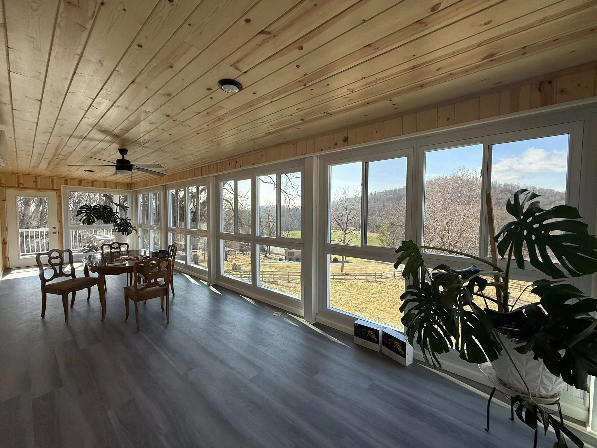 A sunroom with floor-to-ceiling windows showing a view of trees and grass outside. Inside, there are a round dining table with chairs, large indoor plants, a ceiling fan, and a ceiling light, with wood-paneled ceiling and gray flooring.