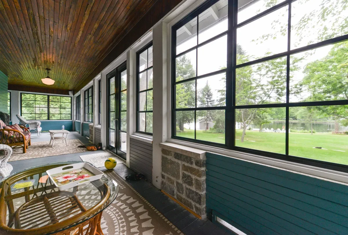 Enclosed porch with large windows showing green outdoor landscape, wooden ceiling, and mix of wicker and wooden furniture.