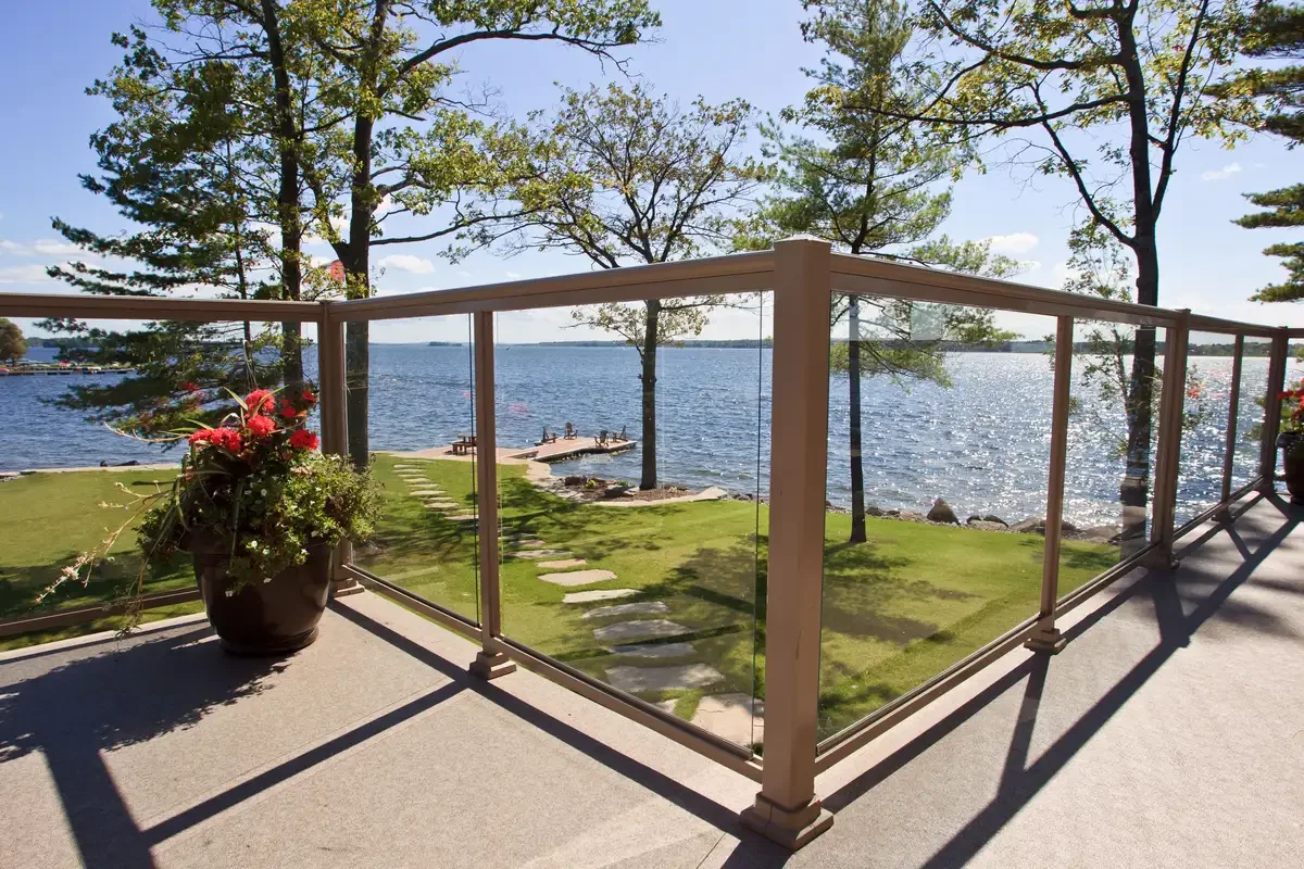 Balcony overlooking a waterfront with trees, grass, a small dock, and a pathway, with a blue sky and sunlight reflecting on the water.