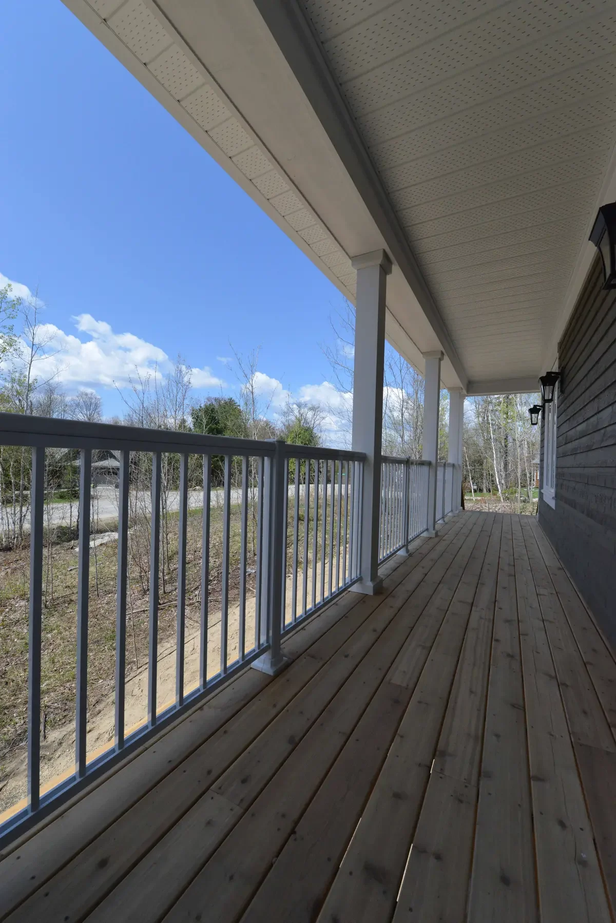 Wooden porch with white railing and support posts, attached to a house with a dark exterior, overlooking trees and a partly cloudy sky.