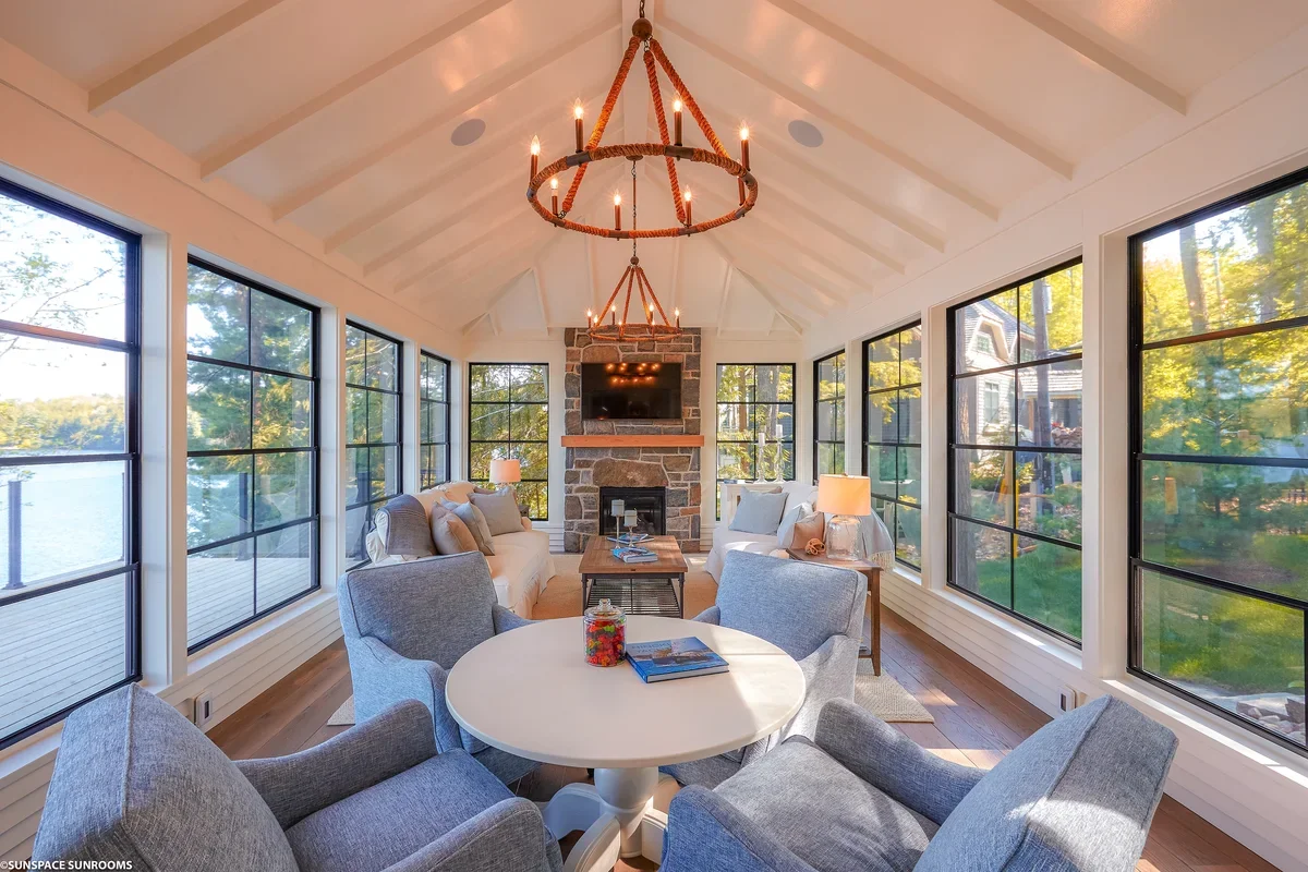 Bright sunroom with large windows showing greenery outside, a stone fireplace with a mounted TV, a round dining table surrounded by gray chairs, and a living area with sofas and side tables illuminated by table lamps and hanging chandeliers.