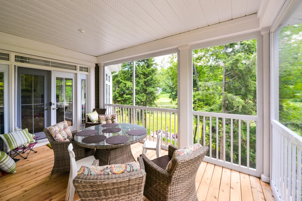 A covered porch or sunroom with a wooden floor, white railings, and several wicker chairs around a glass table, overlooking a green, tree-filled outdoor area.