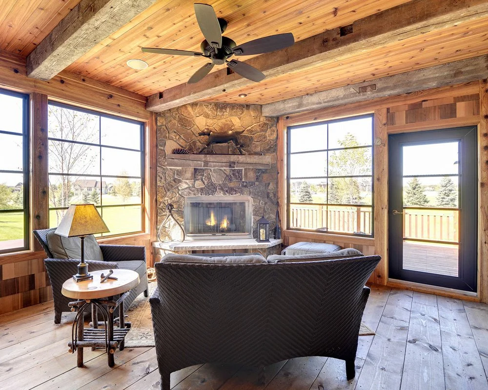 Cozy living room with wood-paneled ceiling, large windows, stone fireplace, ceiling fan, leather sofa, armchair, side table with lamp, and a view of a green outdoor area.