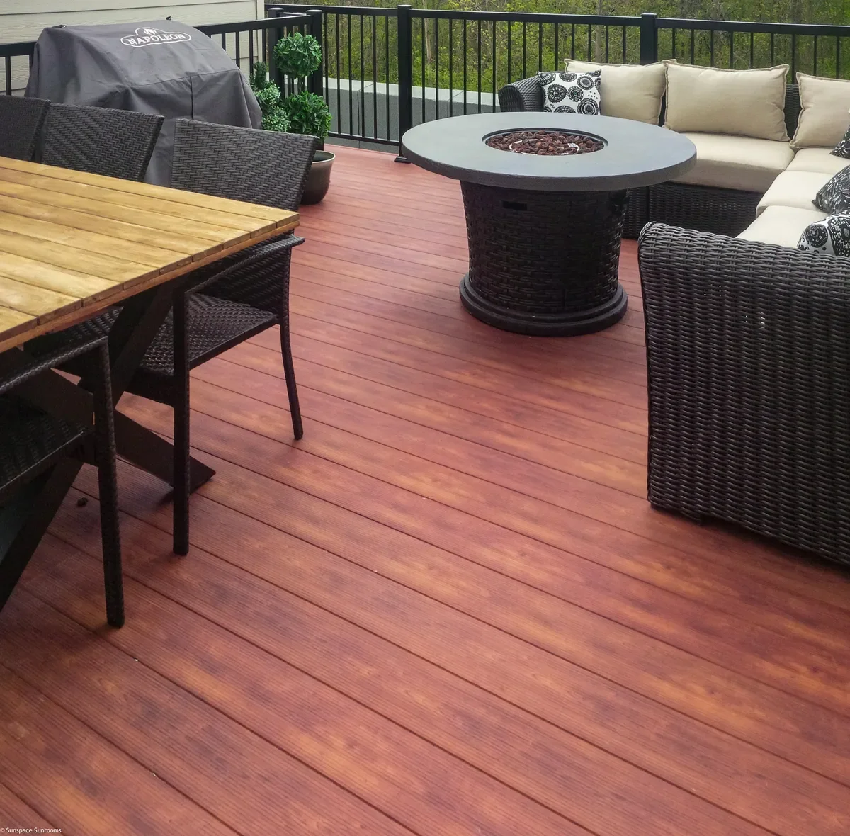 Backyard patio with a wooden dining table, black woven chairs, a round fire pit, a black wicker sectional sofa with beige and black patterned cushions, and a covered grill in the corner, surrounded by a black metal fence.