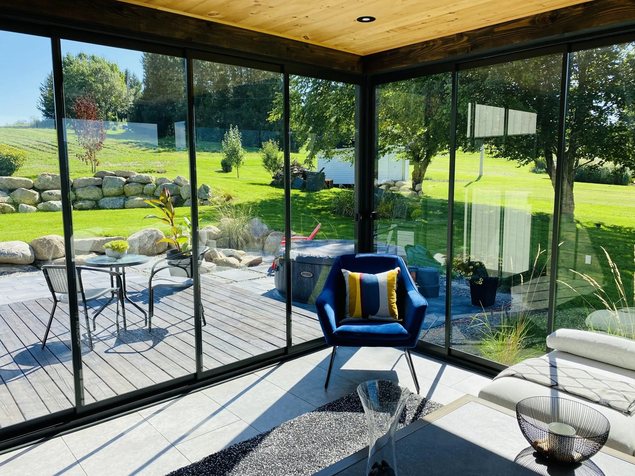View of a lush green yard through glass walls, with rocks, trees, and a small white shed, taken from a modern indoor patio with tiled floor and outdoor furniture.