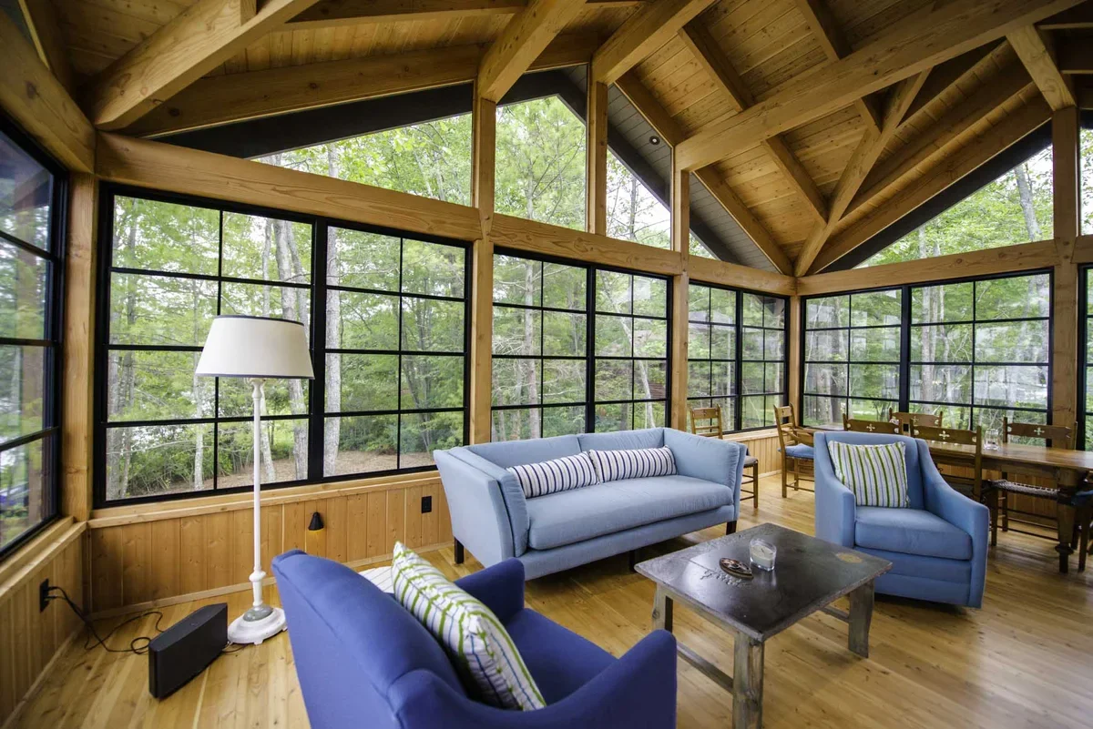 A cozy living room with large windows showing a wooded outdoor view, wooden ceiling and walls, a light blue sofa, blue armchairs with striped pillows, a wooden dining table with chairs, a white floor lamp, and a rustic coffee table.