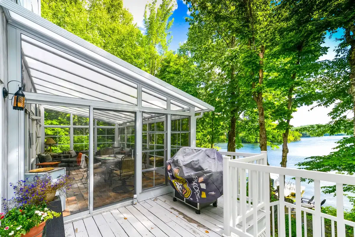 A screened porch with outdoor furniture, potted plants, and a grill, overlooking a river with green trees.