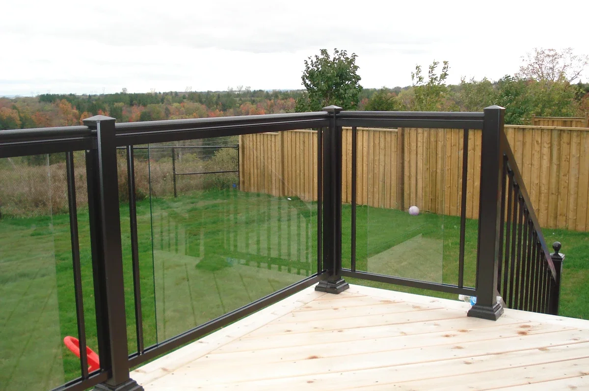 View from a wooden deck with black railing overlooking a backyard with a grassy lawn, a wooden fence, and trees in the distance.