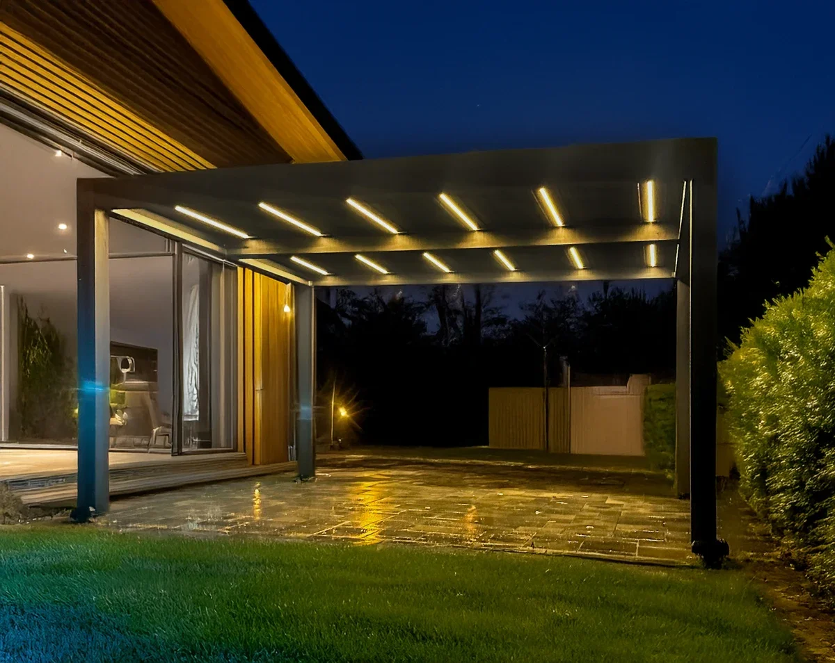 Modern outdoor patio at night with an illuminated metal pergola, glass sliding doors leading into a house, and a grassy yard.