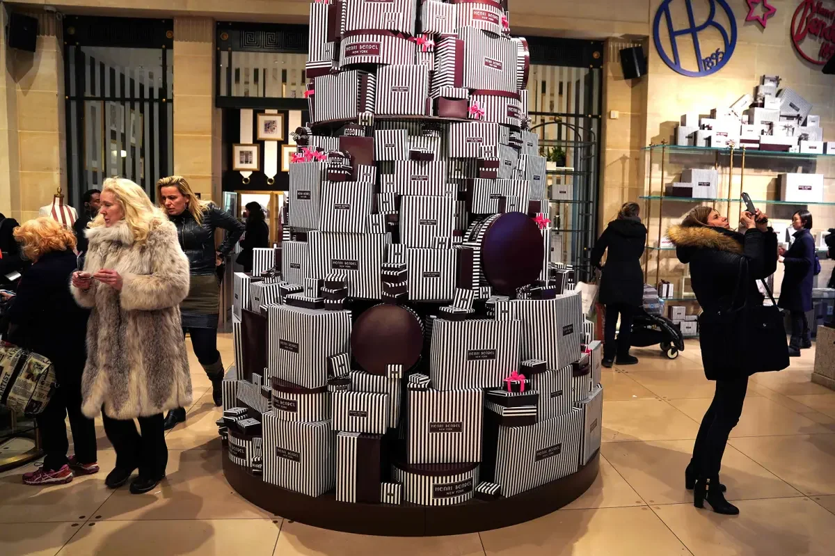 A large Christmas display of stacked black and white striped gift boxes with pink bows, situated in a shopping mall with people shopping and taking photos around it.