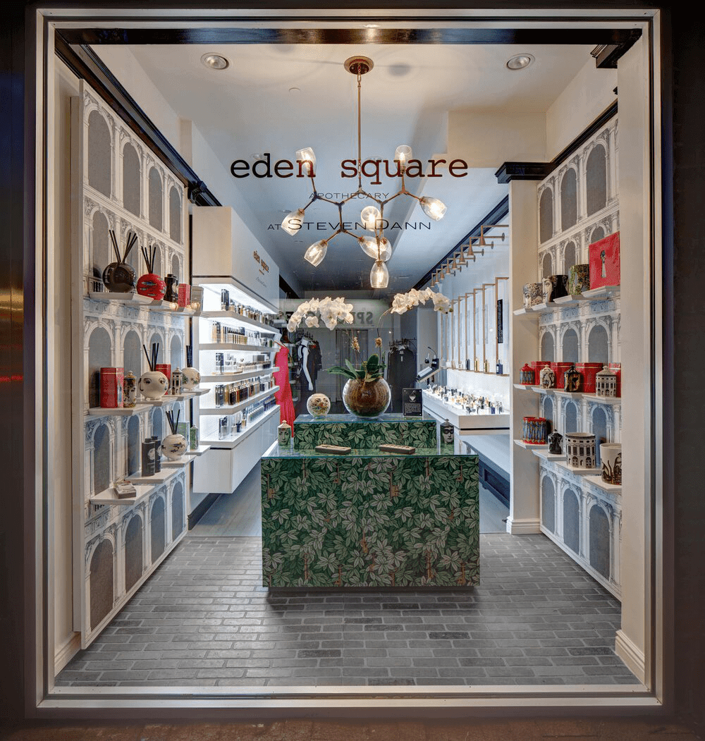 View of a boutique store named Eden Square at Steven Dann, featuring glass display shelves with decorative items and accessories, a green leaf-patterned table with a large plant in a vase, and modern lighting fixtures.