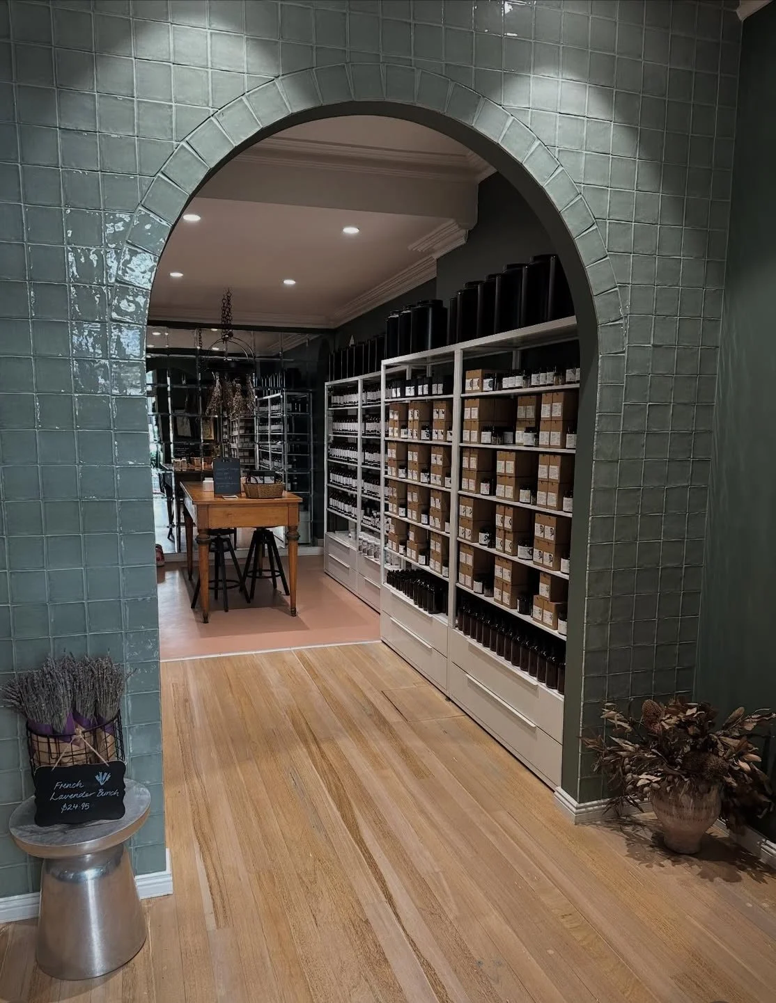 Interior of a boutique store with shelves filled with bottles and boxes, a wooden table with stools, and decorative plants, viewed through a tiled archway.