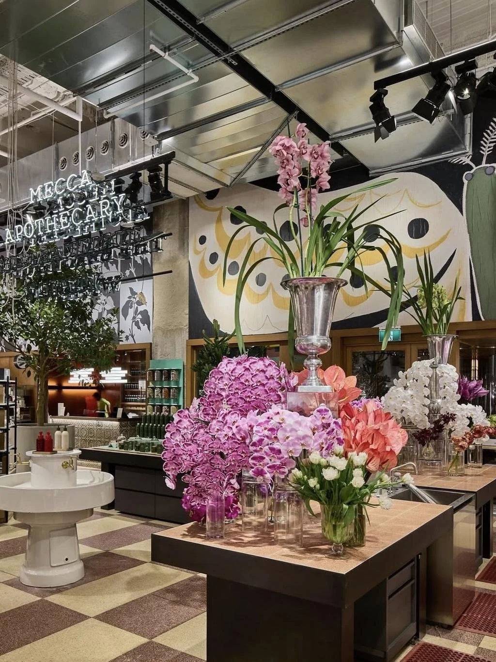 Interior of a store or café with colorful floral arrangements in glass vases on a wooden table, featuring pink, white, and purple flowers. A neon sign reads Scientific & Apothecary, and wall art with abstract designs is visible in the background.