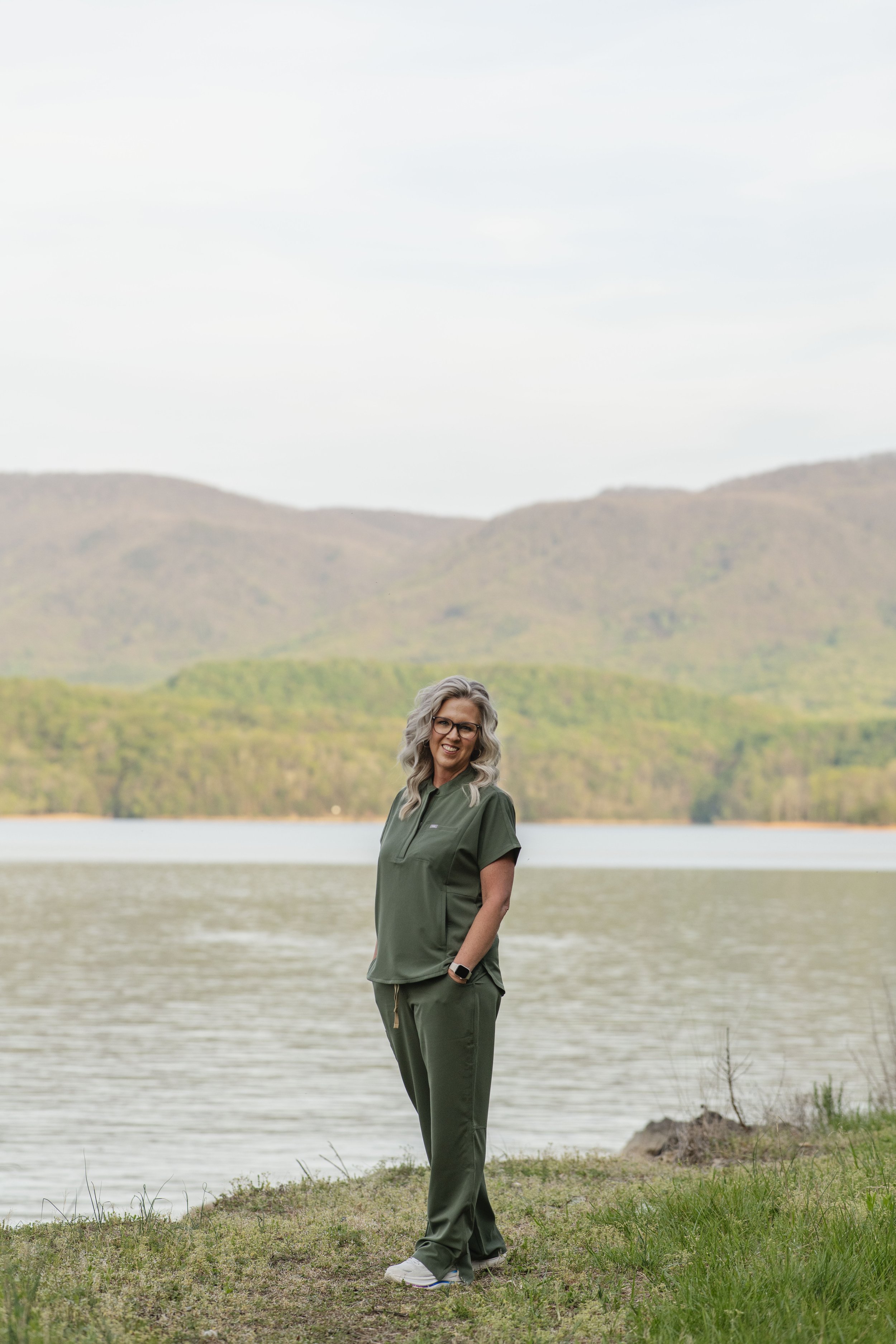 A woman with gray hair and glasses, dressed in a green outdoor outfit, smiling and standing by a lake with hills in the background. SoHo Aesthetics | Botox & Fillers Abingdon VA