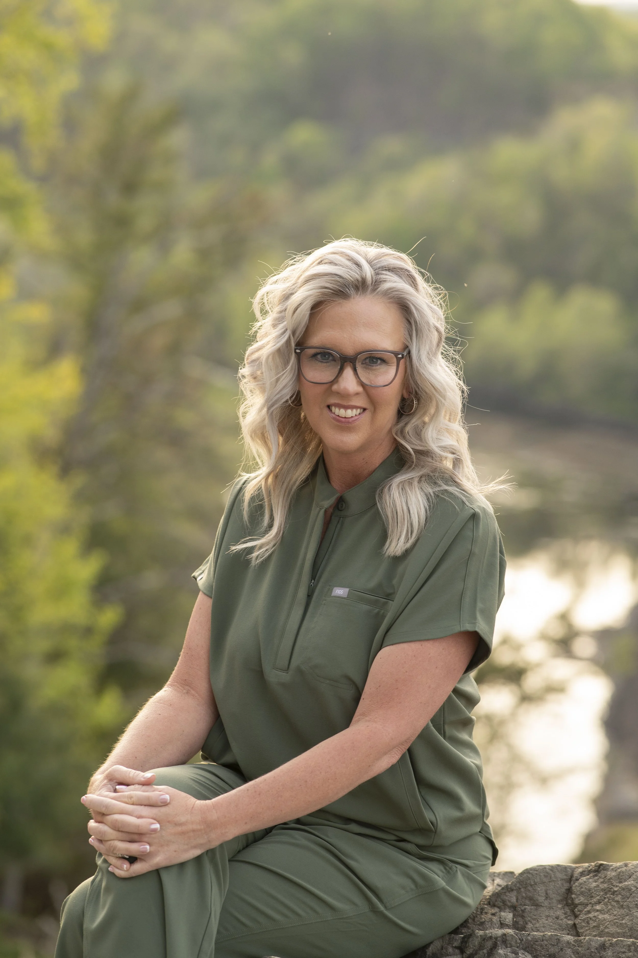 A woman with gray curly hair and glasses sitting outdoors on a rock, wearing a green uniform, smiling at the camera, with a blurred background of trees and water. SoHo Aesthetics | Botox & Fillers Abingdon VA