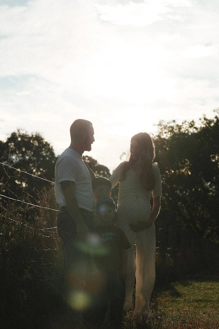 Silhouette of a family of four standing outdoors at sunset, with trees and a fence in the background.