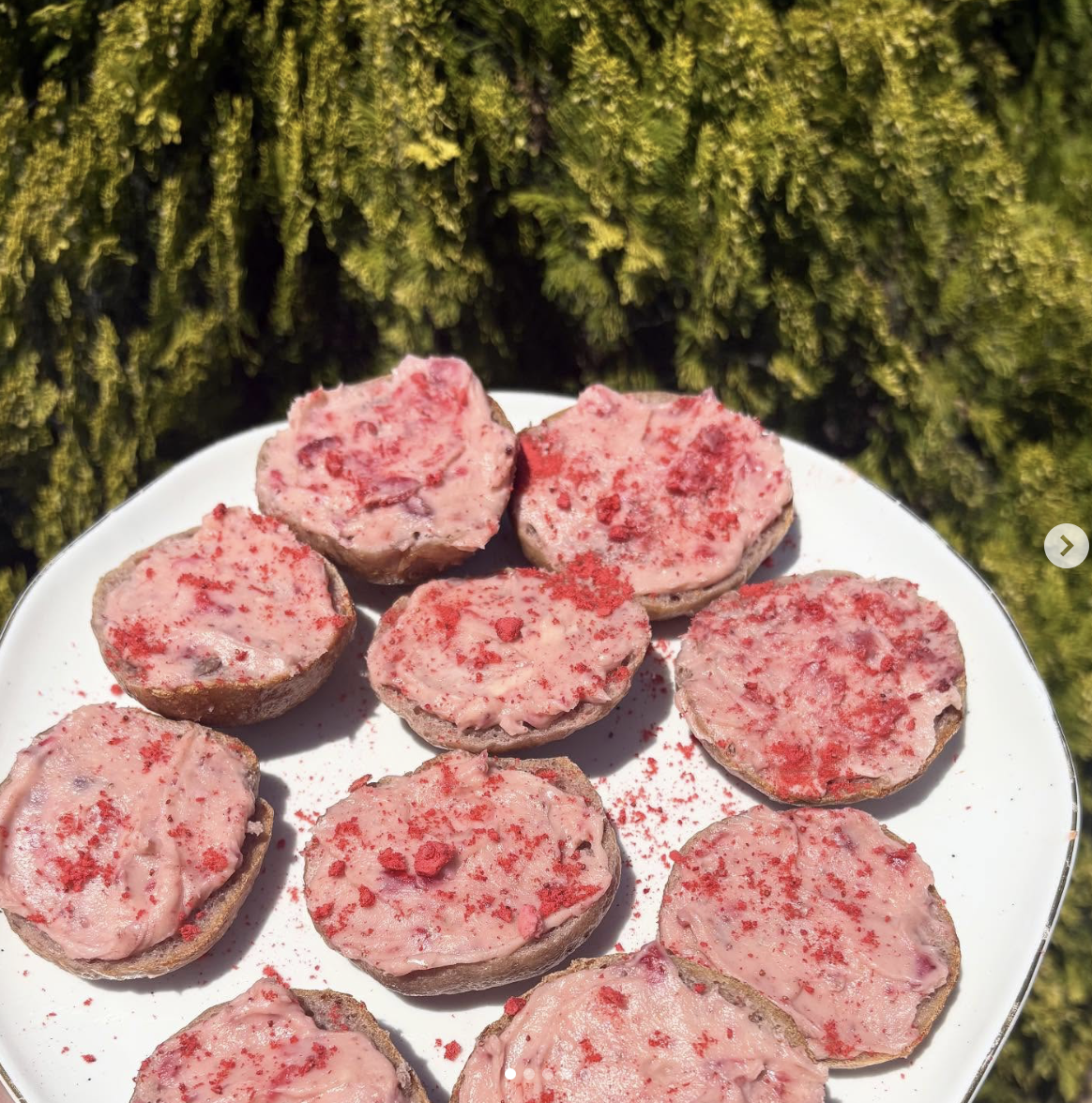 A white plate with several small, round, pinkish-red desserts topped with red powder, set outdoors against a backdrop of green moss or foliage.