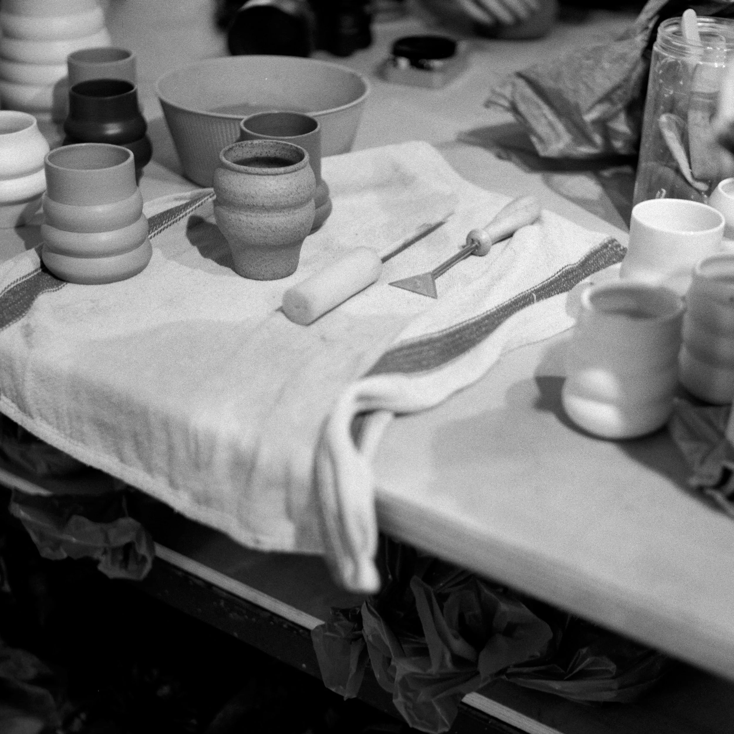 Black and white photograph of a cluttered table with various ceramic cups, a rolling pin, and a plastic container with a lid, along with napkins and other miscellaneous items.