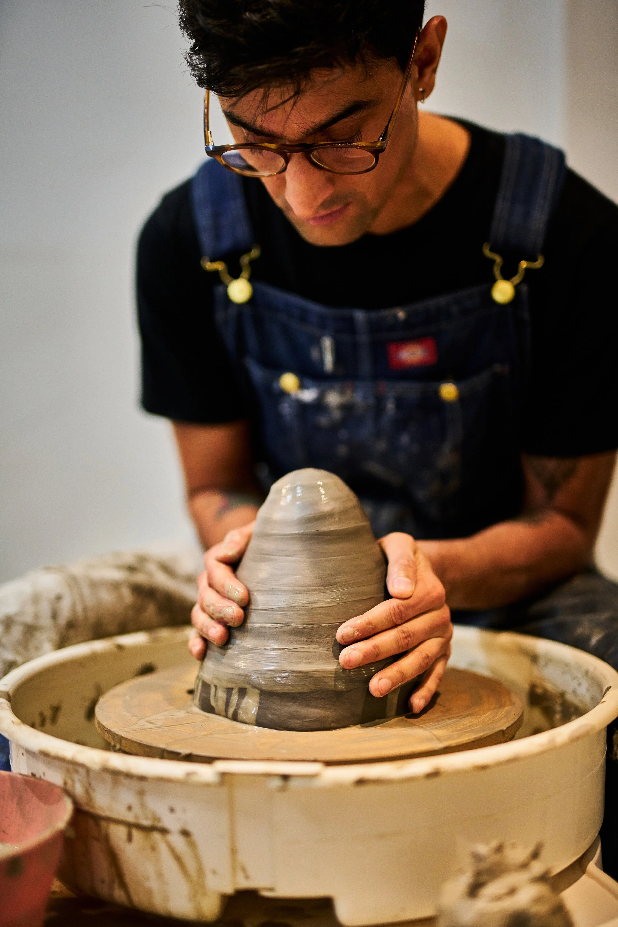 Amar is wearing glasses and a denim apron and s shaping clay on a pottery wheel, concentrating on the task.