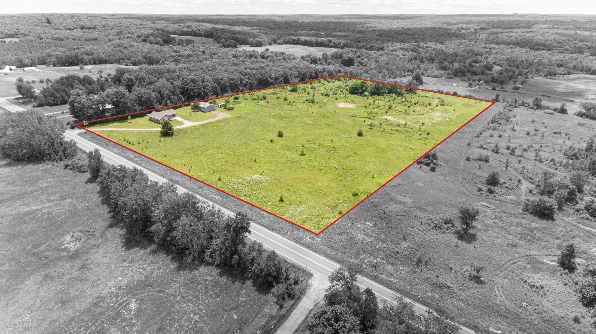 Aerial view of a large plot of land outlined in red, with a small house and outbuildings on one side, surrounded by open fields and trees, with a road at the bottom.