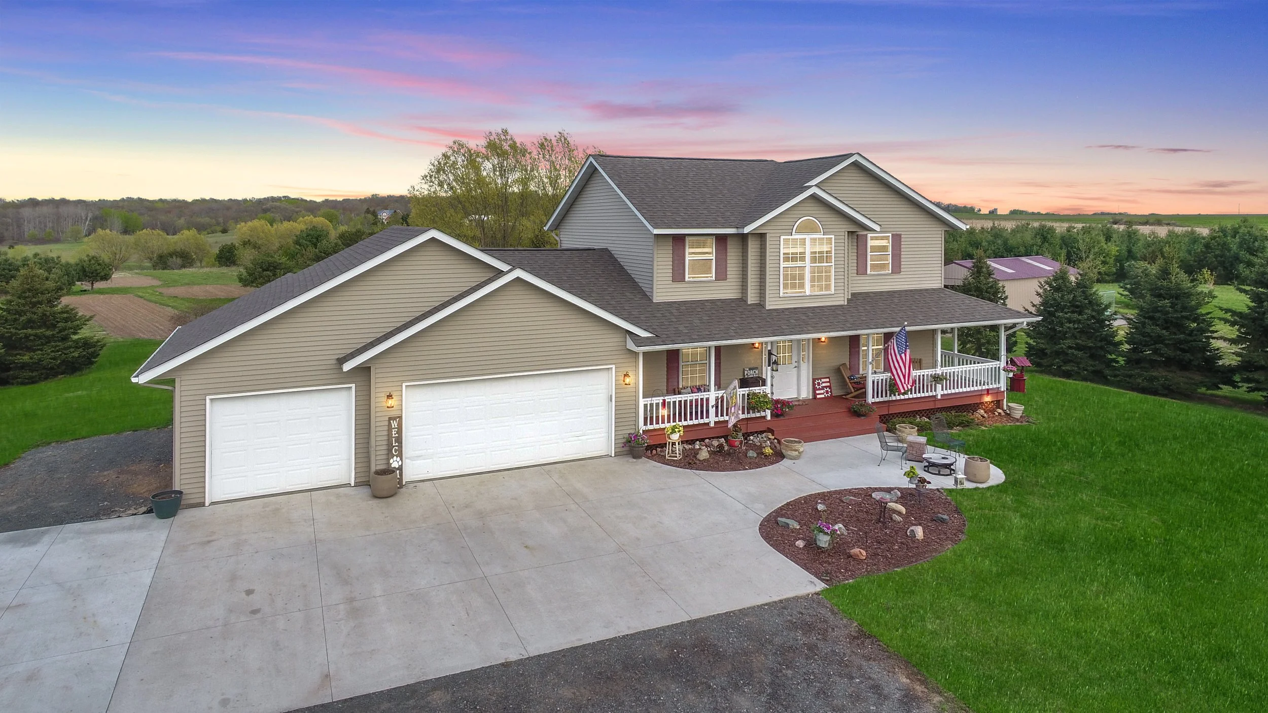 A two-story beige house with white trim, a dark roof, a front porch decorated with an American flag, potted plants, and outdoor seating, set against a rural landscape at sunset.