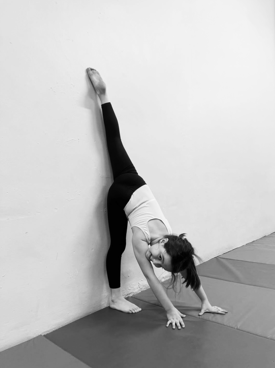 A girl practicing yoga against a white wall, performing a downward dog pose with one leg extended upward and her head near the ground.