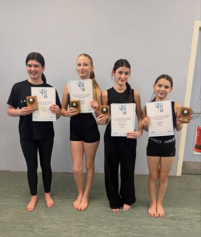 Four young girls standing indoors, holding certificates and small wooden plaques, smiling for a photo after a gymnastics or dance event.