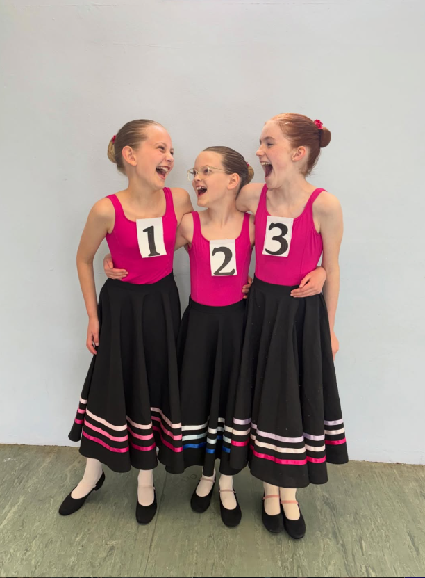 Three young girls in matching pink dance costumes and black skirts, numbered 1, 2, and 3, standing together and laughing against a plain white wall.