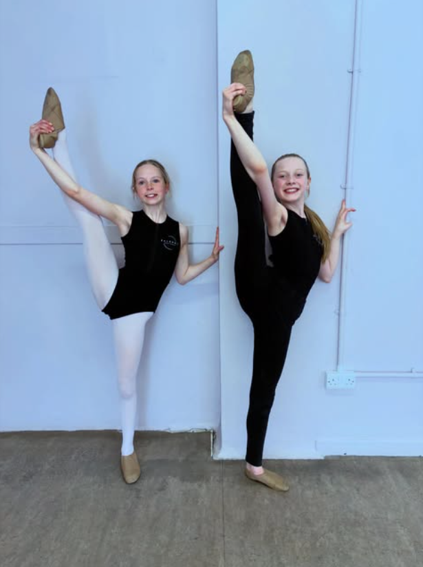 Two young girls in ballet attire performing flexibility poses against a white wall in a dance studio, each balancing on one leg with the other leg raised high.