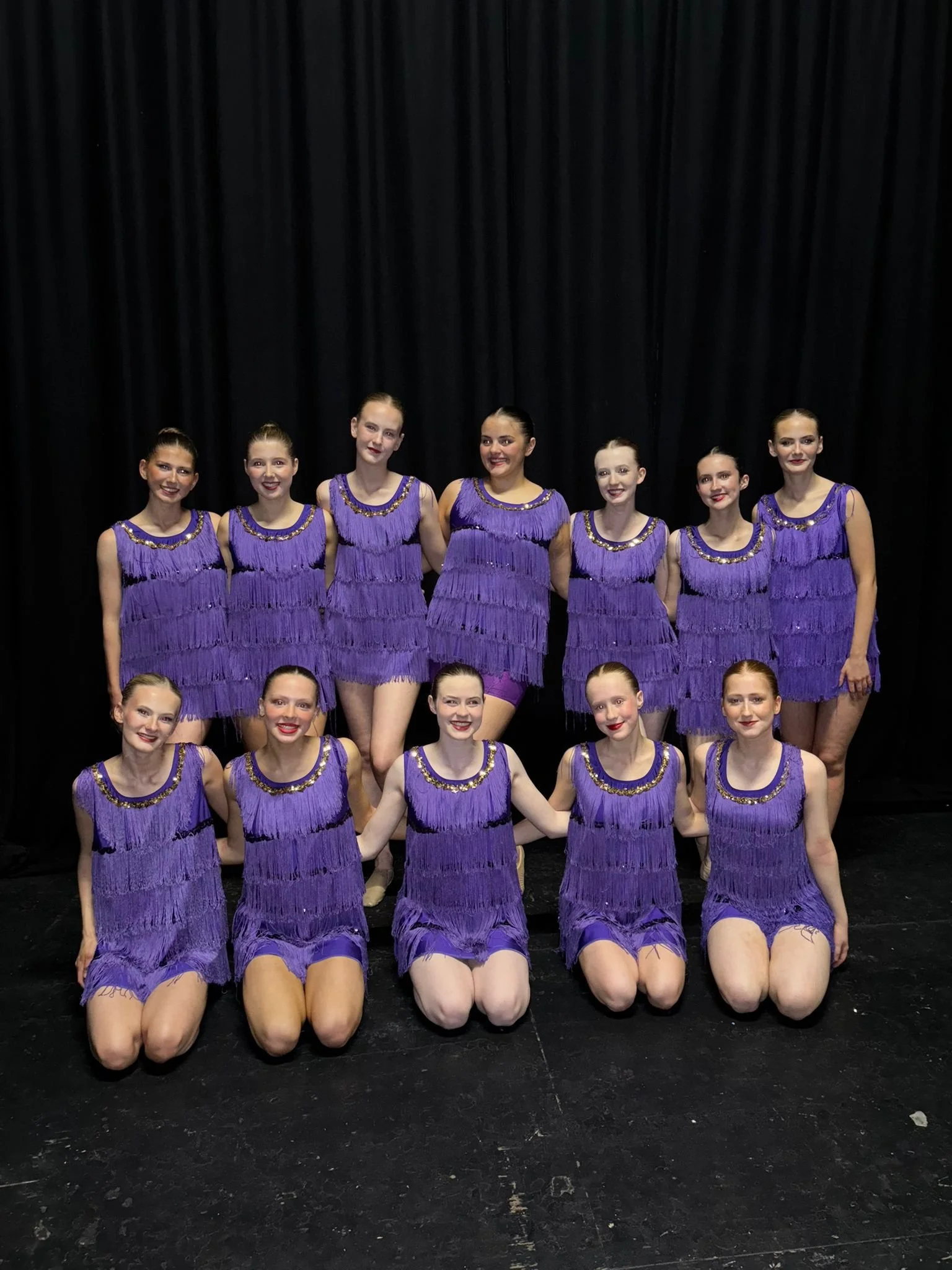 Group of young female dancers dressed in purple costumes, posing on a stage with a black curtain backdrop.